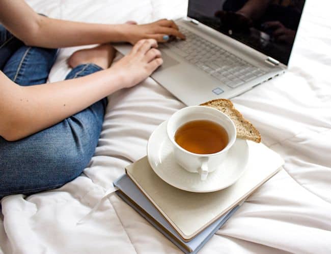 Girl using laptop sitting on white blanket on the bed.  This photo is used for The Frugal Housewife's Contact Us page.  
