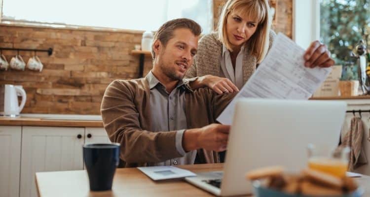 Couple looking over the bills. Man is sitting at desk and woman is standing behind him looking at a bill.