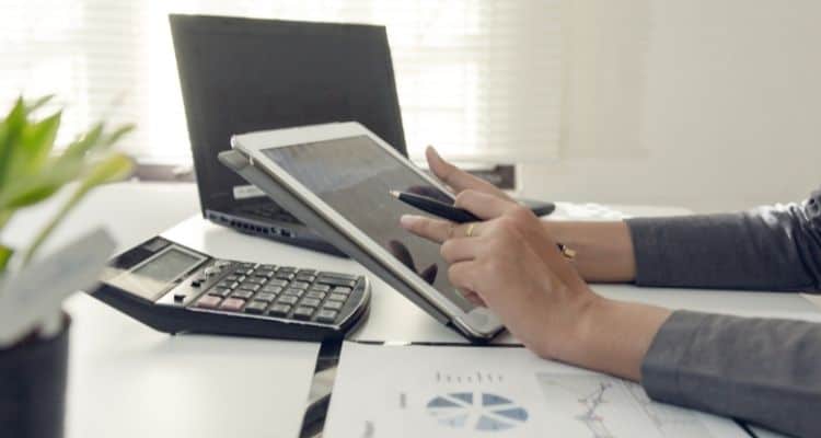 Woman using a tablet at a desk with a calculator and laptop sitting on the desk.
